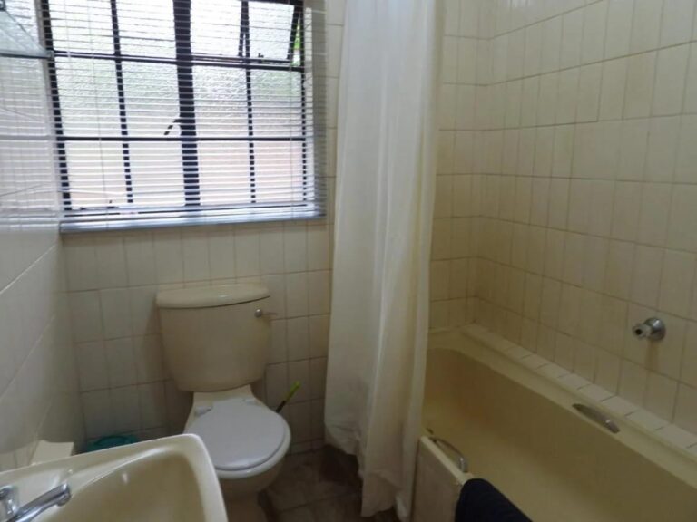 Een betegelde badkamer met een bad met douchegordijn, een toilet en een wastafel, onder een raam met rolgordijn