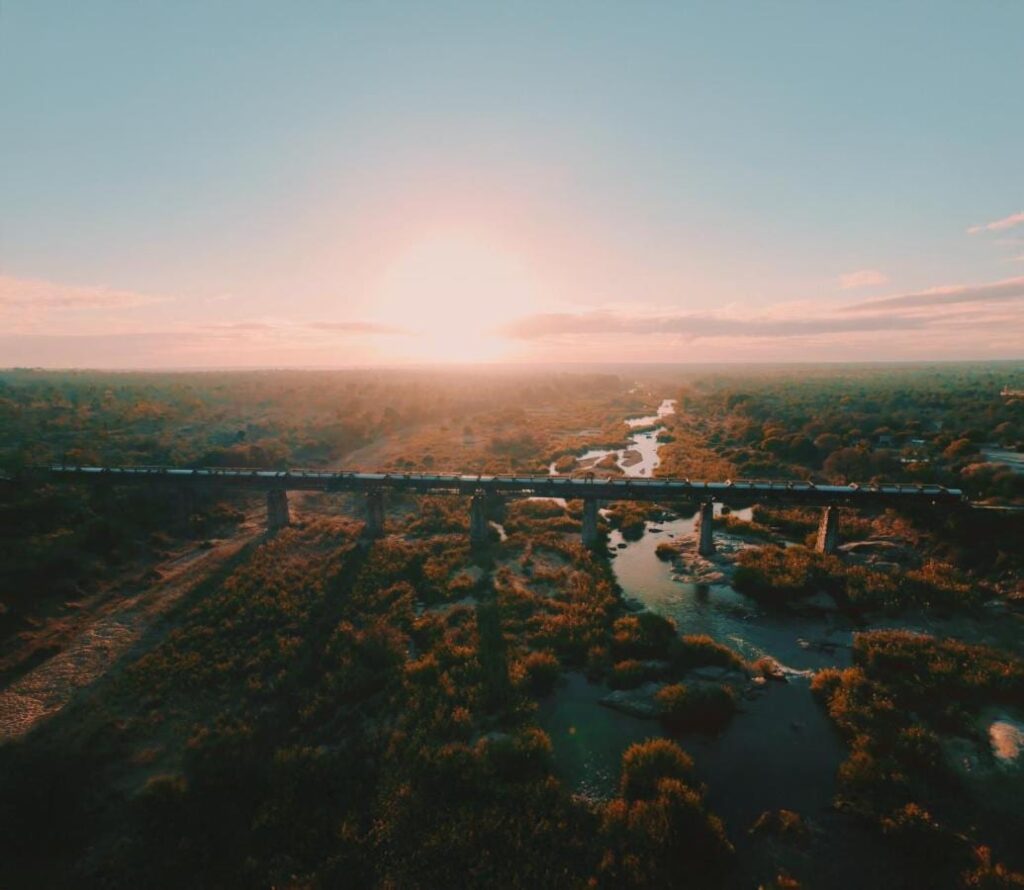De trein van Kruger Shalati op de Selatibrug, van bovenaf gezien, boven de rivier en de bush, met de zon laag aan de horizon