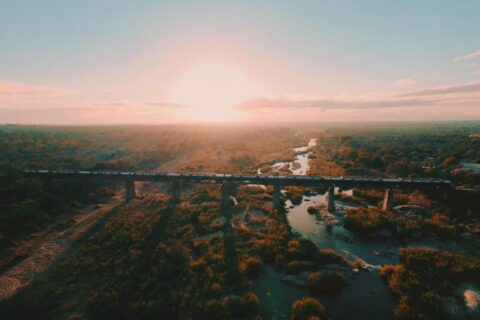 De trein van Kruger Shalati op de Selatibrug, van bovenaf gezien, boven de rivier en de bush, met de zon laag aan de horizon