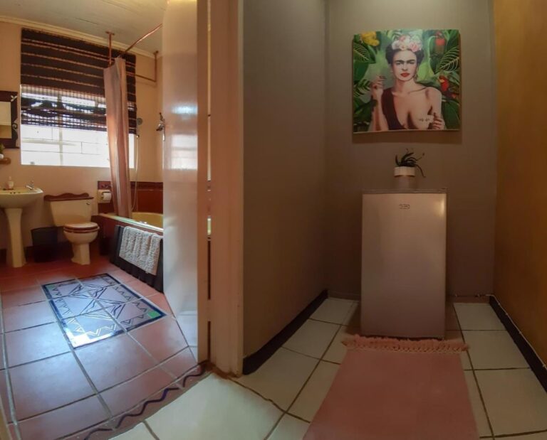 Een badkamer met bad, wastafel en toilet, gezien vanuit een gang waar een kleine koelkast onder een vrouwenportret staat