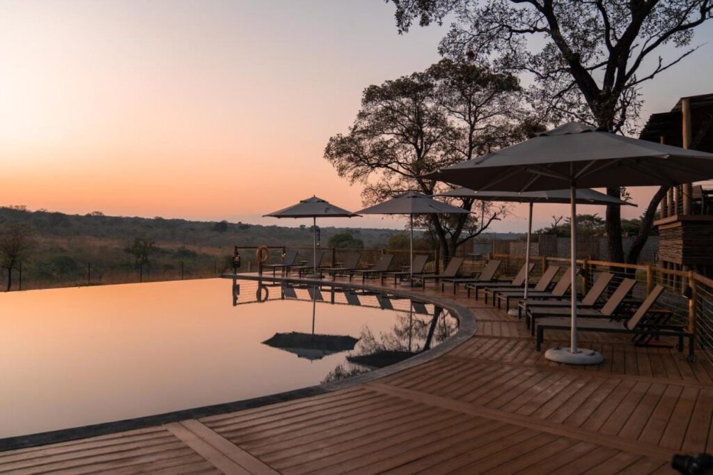 Het infinityzwembad van de Mdluli Safari Lodge onder een oranje lucht, met ligbedden en parasols eromheen