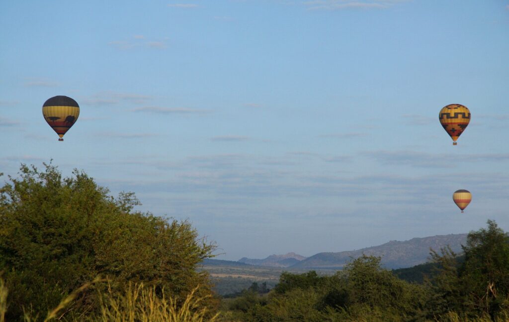 Drie geel met zwarte heteluchtballonnen zweven in een lichtblauwe lucht boven de groene bush, met de heuvels van de krater aan de horizon