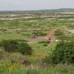 Uitzicht vanaf Observation Hill over een groene vlakte met zoutvlekken, een rode weg, drie 4x4's geparkeerd bij een klein okerkleurig gebouw, lage heuvels in de verte