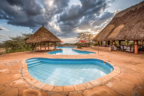 Een klein betegeld bad op de voorgrond en het grote zwembad van de Samburu Sopa Lodge, tussen een strohut en een terras met rieten dak