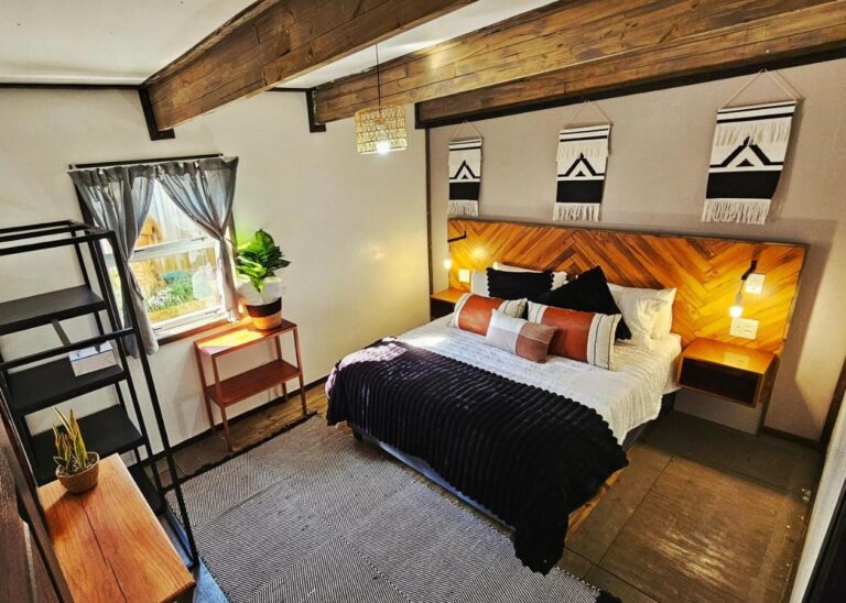 Een kamer in The Elements met een tweepersoonsbed, een houten hoofdeinde in visgraatmotief en drie zwart-witte wandkleden, onder zichtbare balken