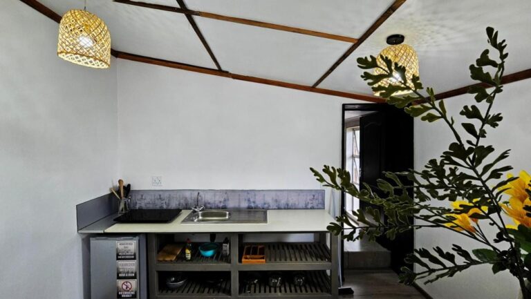 Een kitchenette met kookplaat, gootsteen, kleine koelkast en open planken, onder twee rieten hanglampen
