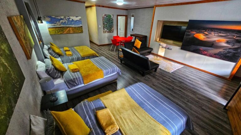 Een grote accommodatie met drie bedden met gele plaids, een zwarte bank, een rode tafel en grote landschapsfoto's aan de muren