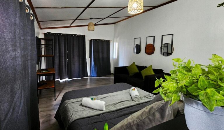 Een grijs tweepersoonsbed met opgerolde handdoeken, een zwarte bank en donkere gordijnen, in een studio met een houten rasterplafond