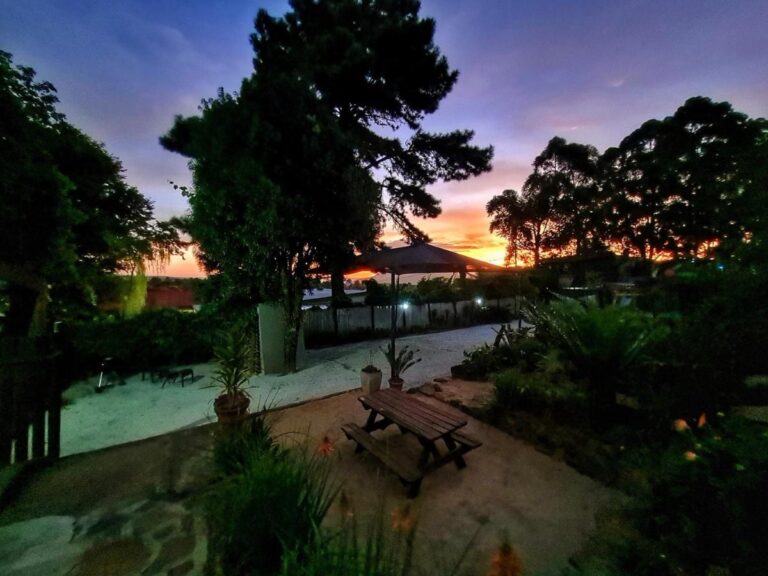 De tuin bij zonsondergang, met een picknicktafel onder hoge bomen en een oranje lucht