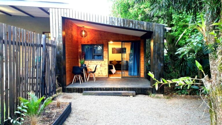 Een houten chalet met een overdekt terras, een tafel en twee zwarte stoelen, voor een binnenplaats met grind en planten