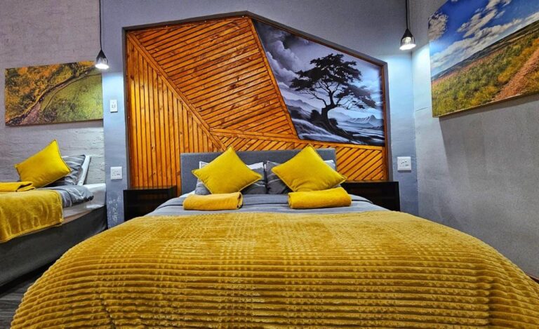 Een tweepersoonsbed met een gele plaid voor een hoofdeinde van houten latten en een zwart-wit schilderij van een boom, naast een tweede bed